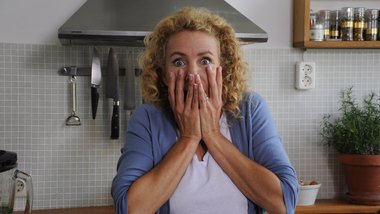 Eine Frau in einer Küche blickt mit beiden Händen im Gesicht verstört in den Raum.